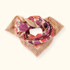 Grand foulard "Latika Joy Macadamia" de la marque Apaches Collections. Accessoire en coton doux avec un imprimé floral aux teintes vives de rouge, violet et orange, sublimé par des bordures beige rayées. Ce foulard polyvalent et tendance peut se porter en bandana, autour du cou ou comme accessoire de cheveux pour un style bohème et élégant.